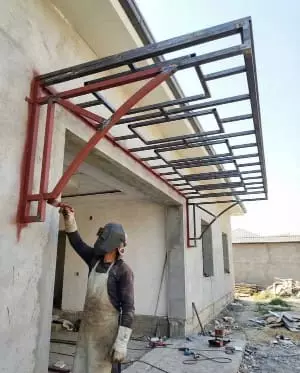 A worker wearing a welding mask and apron installing a metal canopy
