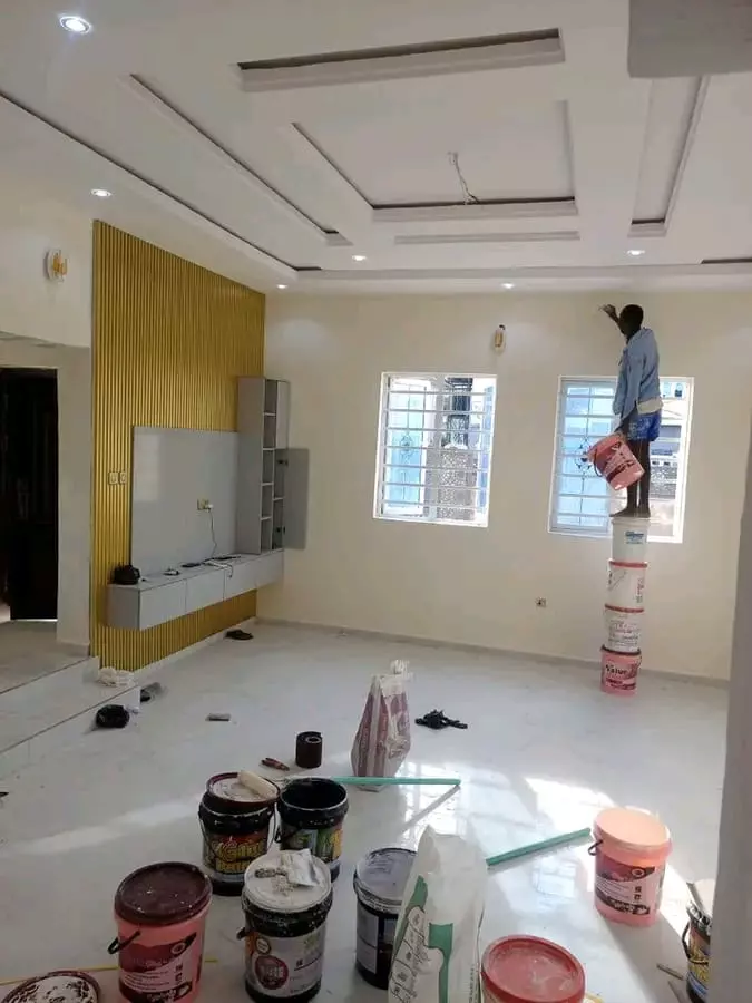 A worker balancing on a stack of paint buckets to paint an interior wall