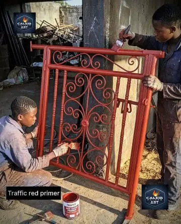 Two workers painting an ornate red metal gate with primer